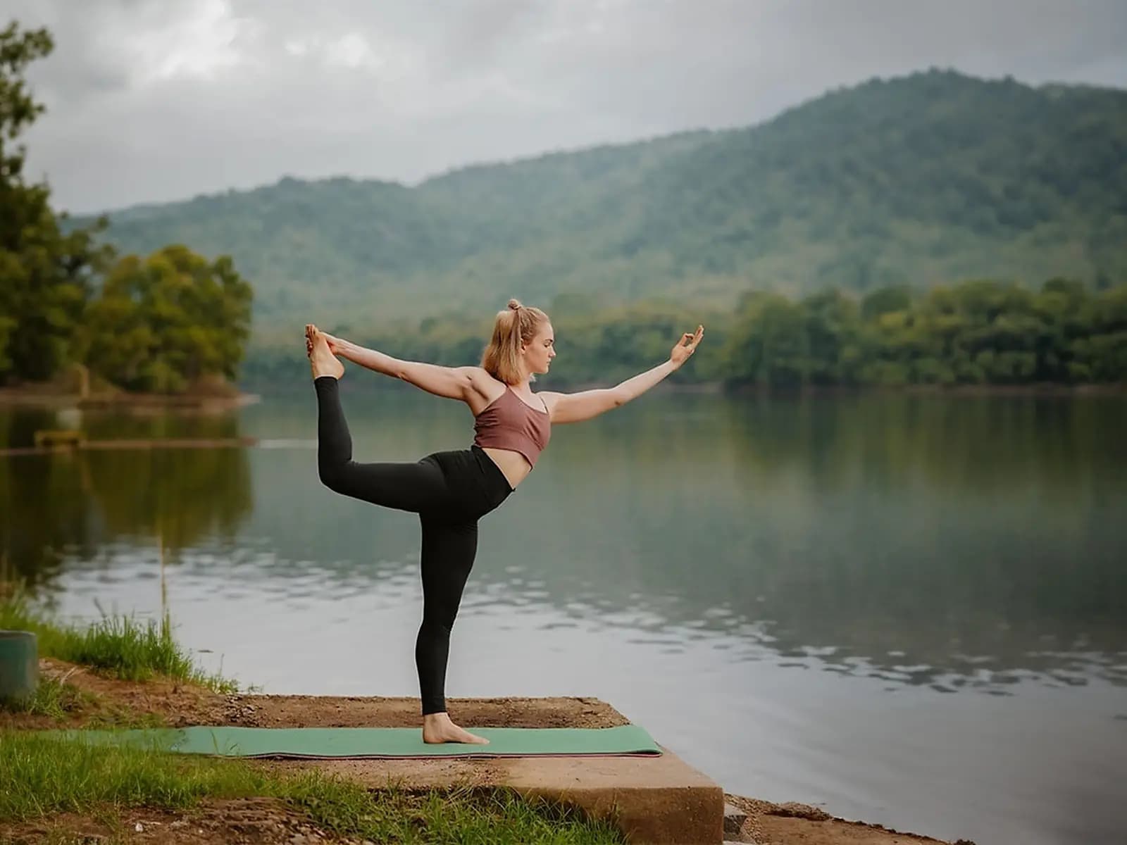 Woman in a standing Yoga balance on a stone ledge by a river below green hills in Kerala