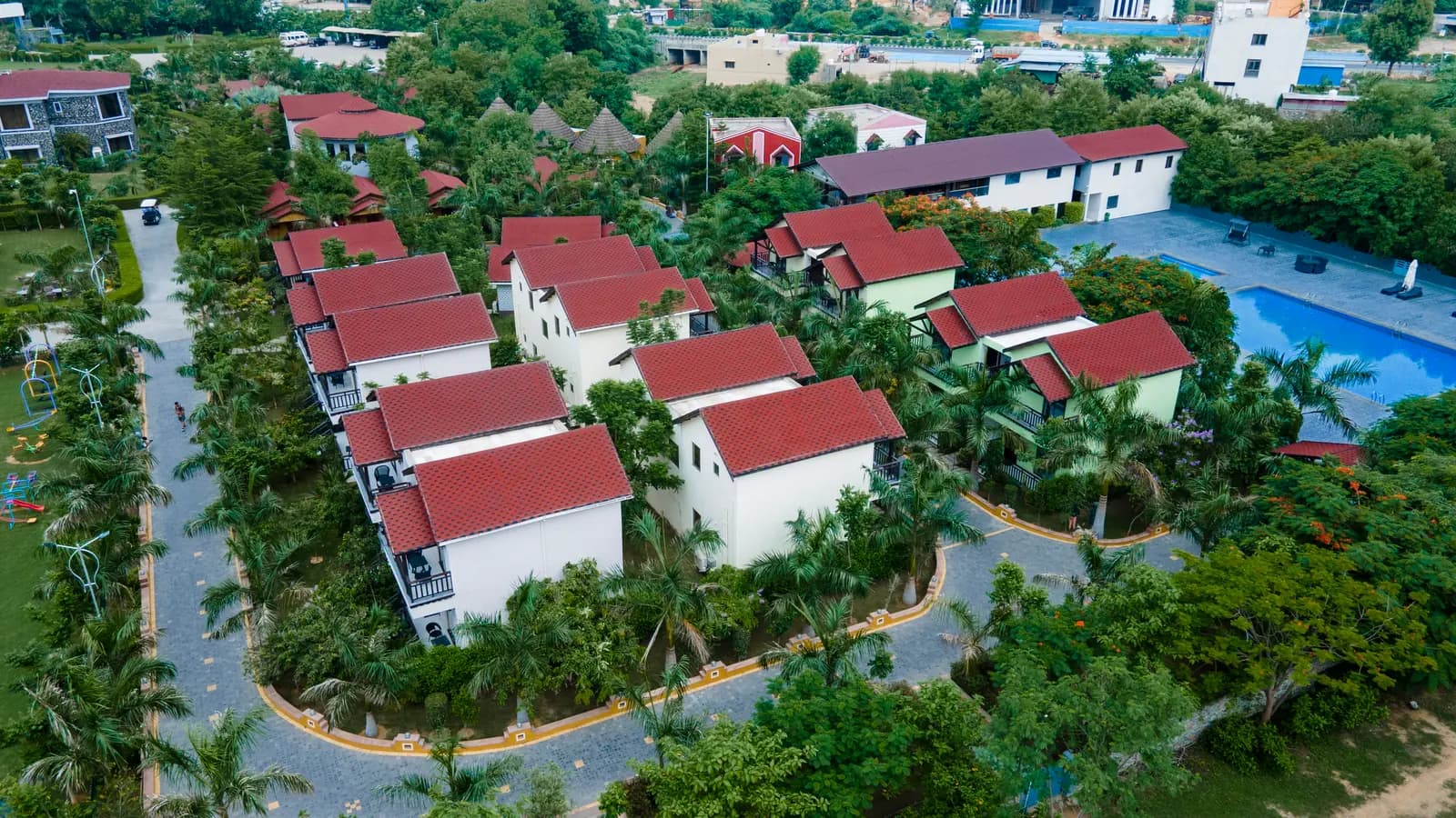 Aerial view of white cottages with red roofs and a pool among palms at an Ayurveda resort in Abu Road